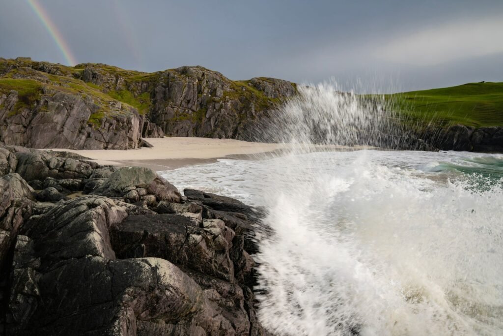 Waves breaking below a rugged Scottish coastline beneath a rainbow.