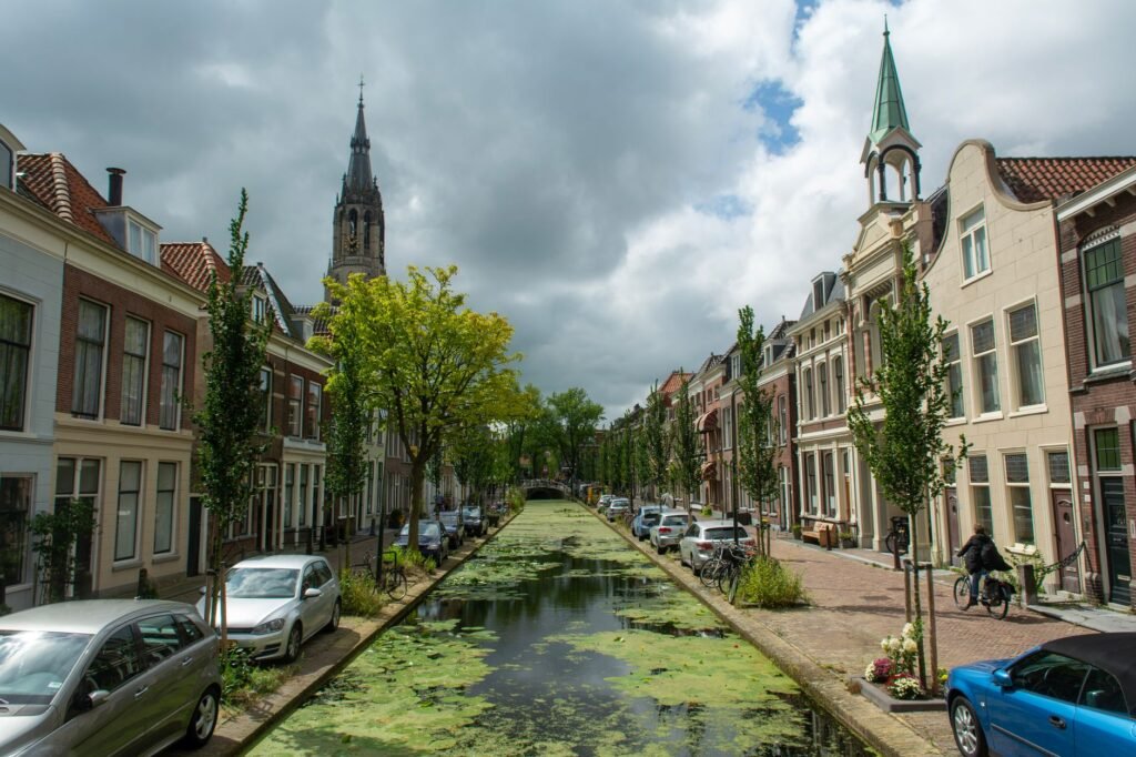 A calm Delft canal bordered by trees, brick houses and parked bicycles beneath a cloudy sky.