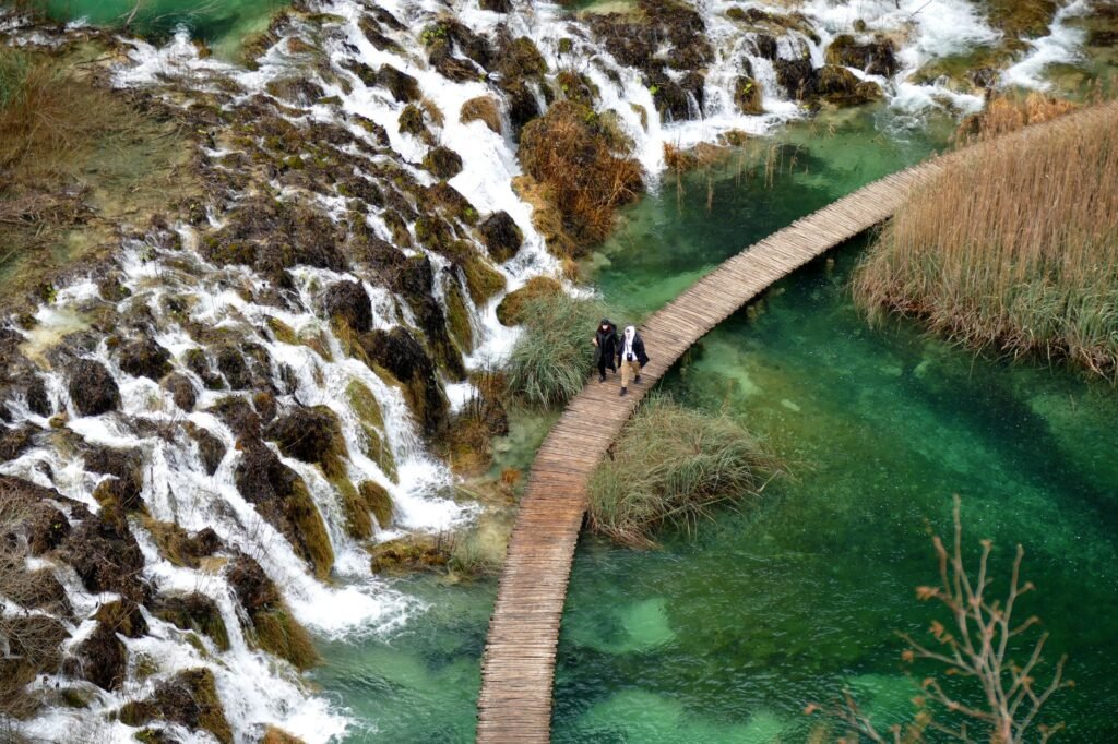 Two visitors walking on a narrow wooden boardwalk above green water at Plitvice Lakes in Croatia.
