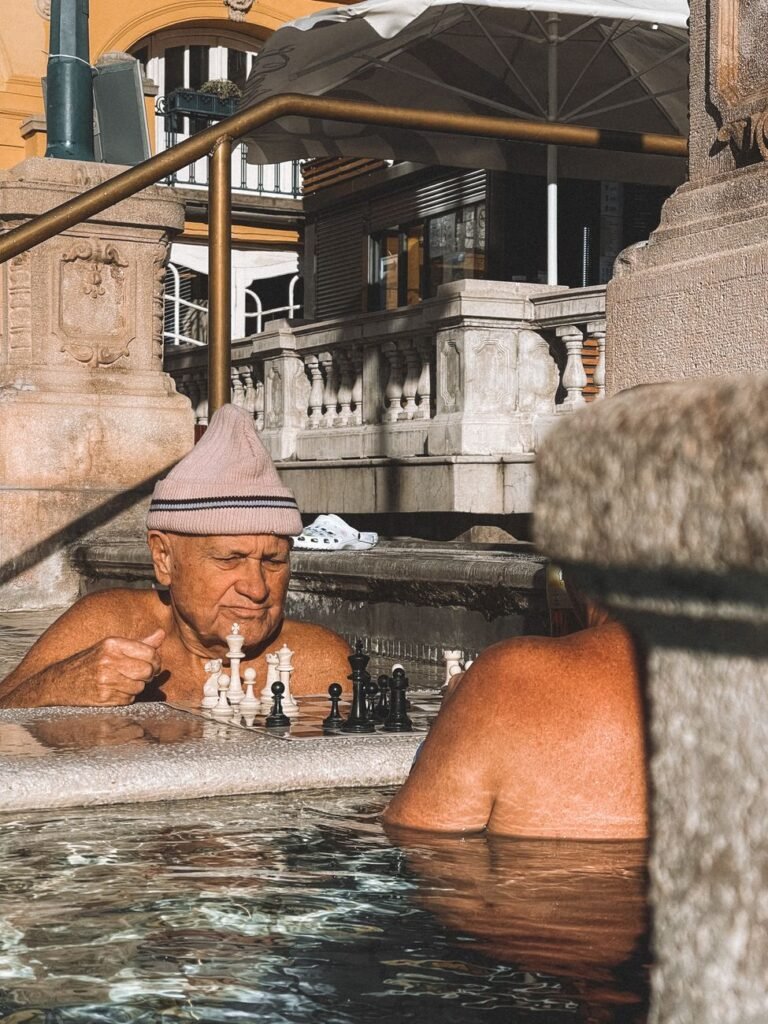 Two older men playing chess on a floating board while standing in a thermal pool in Budapest.