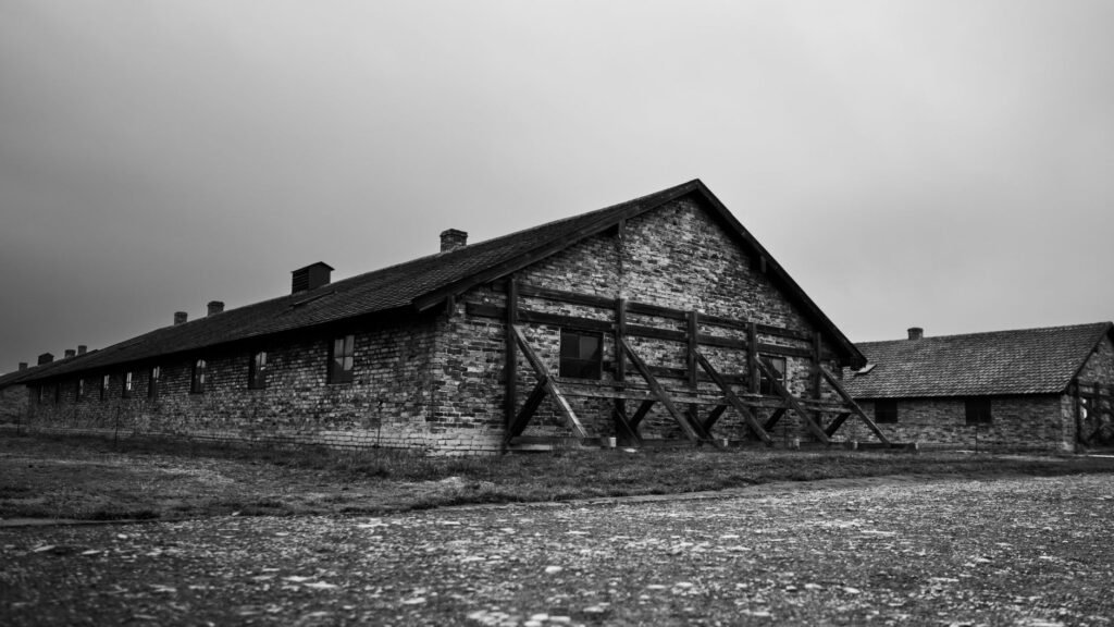 Black-and-white view of wooden barracks and fencing beneath clouds at Auschwitz II-Birkenau.